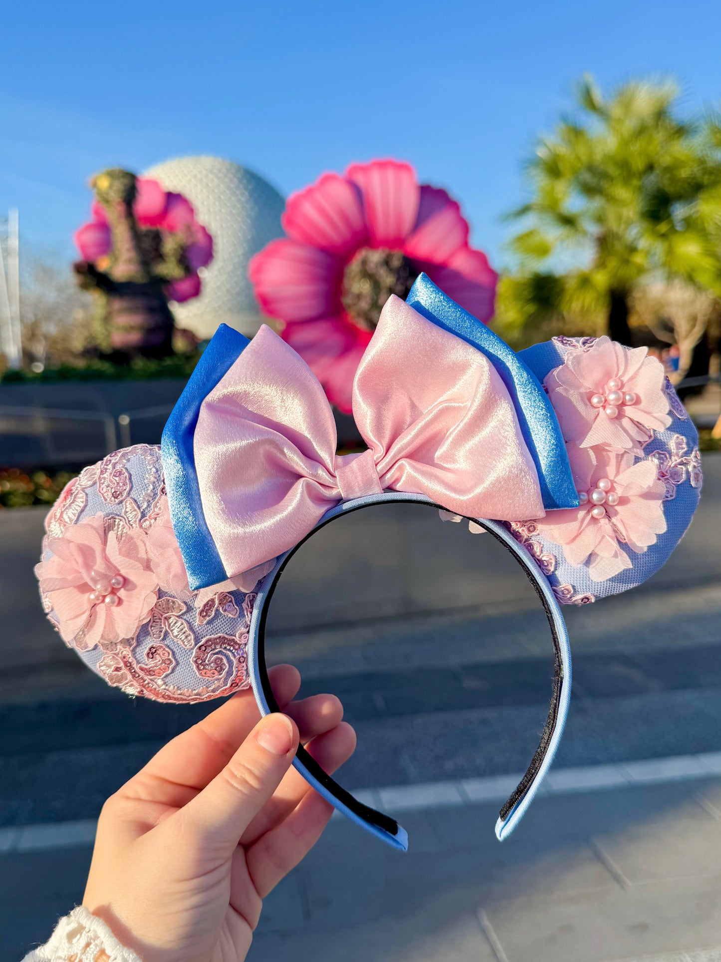 pink floral ears being held in front of some flowers and Soaceship Earth at Epcot