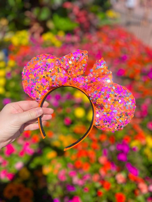 Hand holding orange, pink, and silver sequin mouse ears in front of flowers of the same colors.