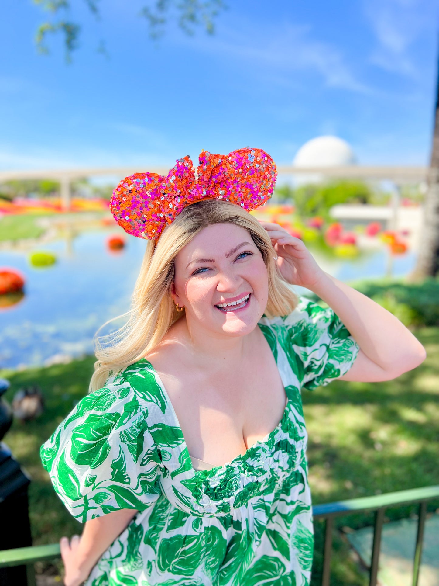 A person wearing the sequin mouse ears in front of Epcot wearing a green and white floral dress.