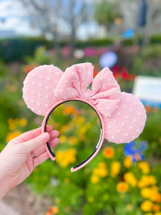 A pink/peach pair of mouse ears with swiss dots displayed in front of a floral background.
