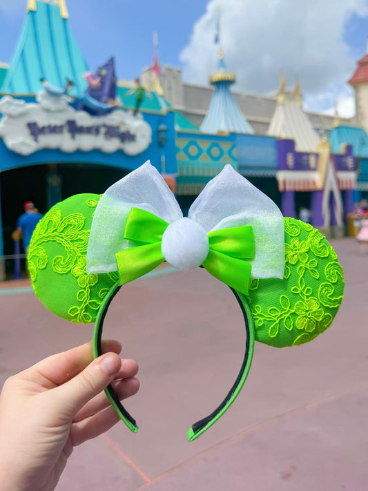 Green and white lace mouse ears with white bow, green ribbon, and white pom held in front of a Fantasyland background