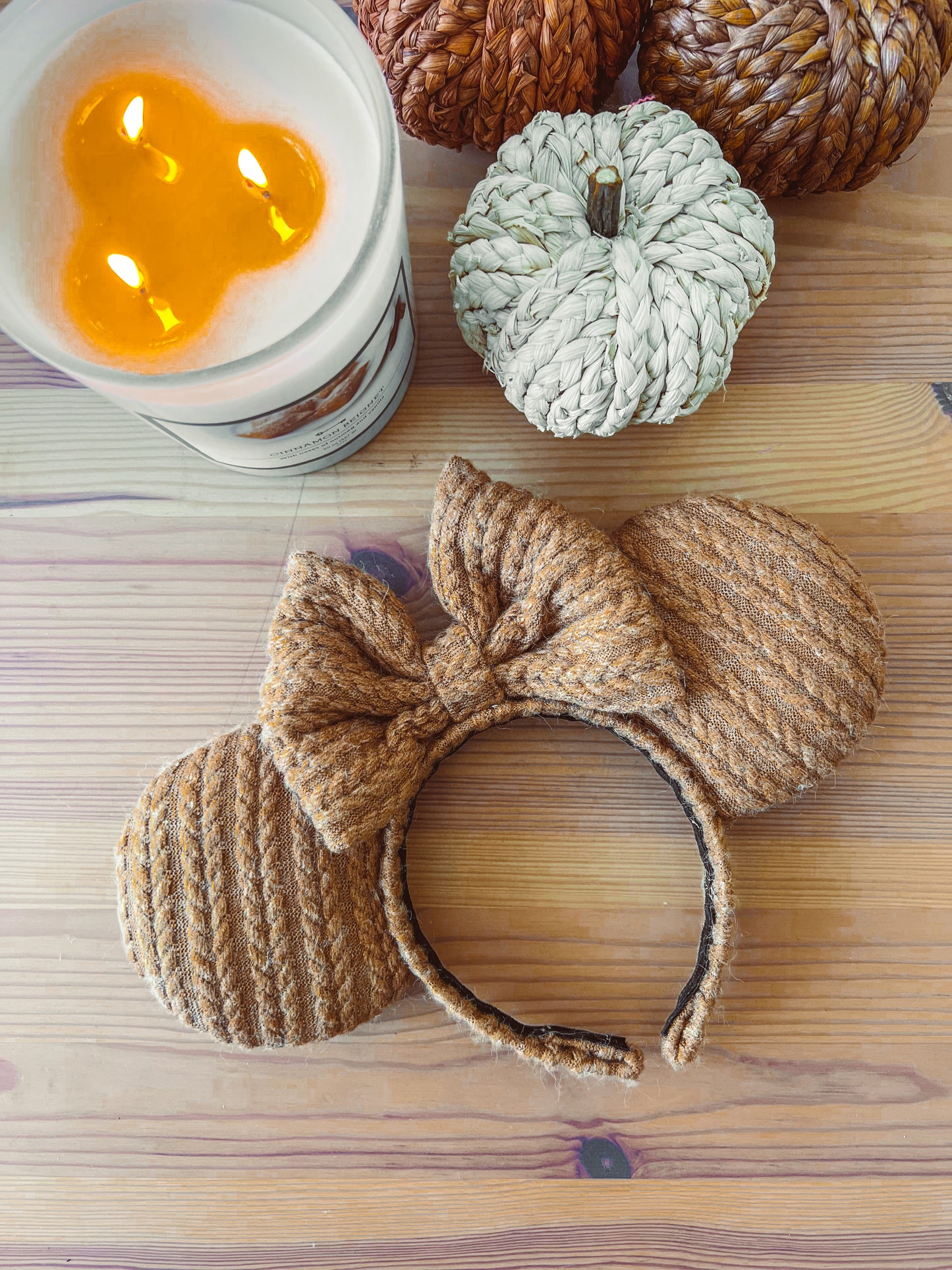 A pair of caramel acorn colored cable knit ears with a large bow, displayed next to a white pumpkin and a candle.