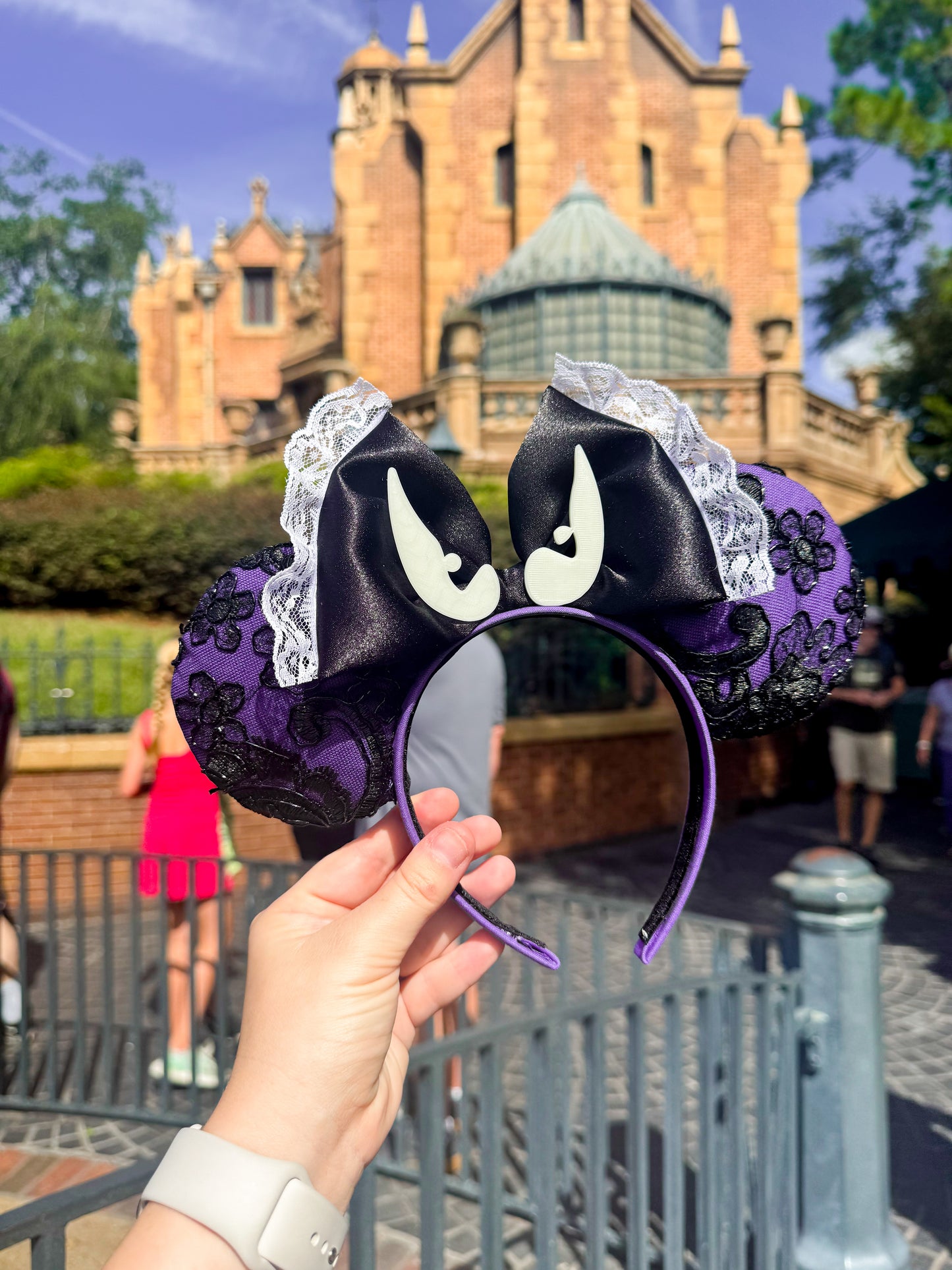 Purple and black lace ears with a black bow and glow in the dark bat eyes on it. The background is a theme park.