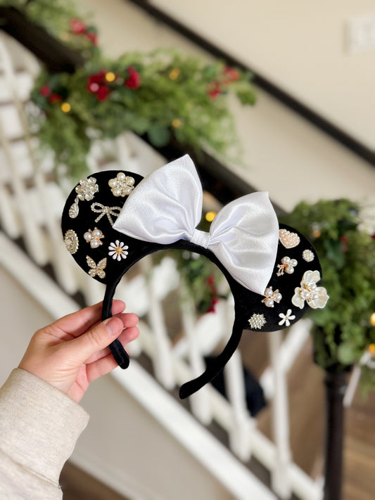 Black velvet mouse ears with white bow and floral pearl and rhinestone embellishments held by a hand, with a blurred decorative background.