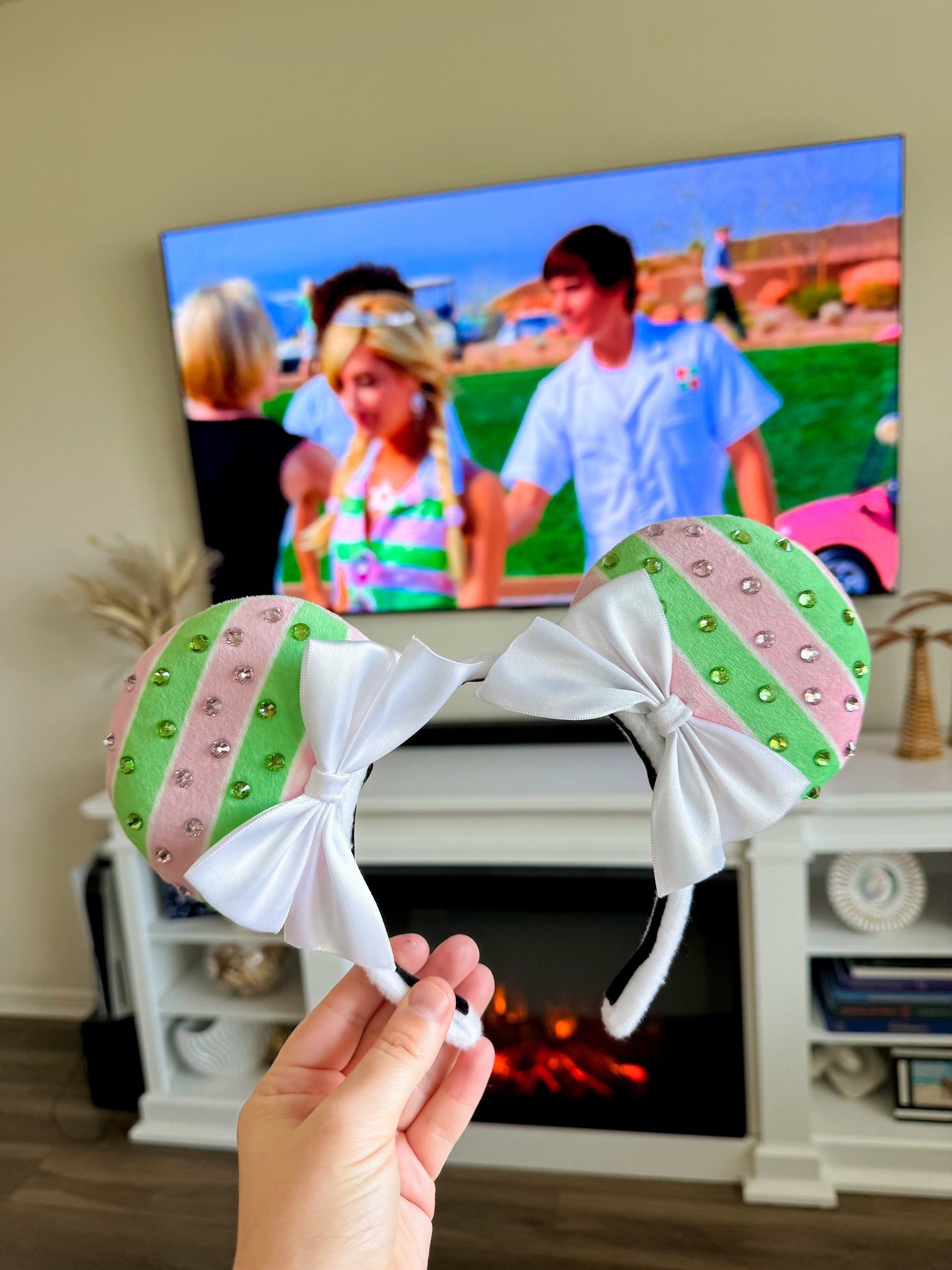 A hand holding a pair of fabric ears with a striped pattern in front of a TV showing a scene from High School Musical with Sharpay Evans.