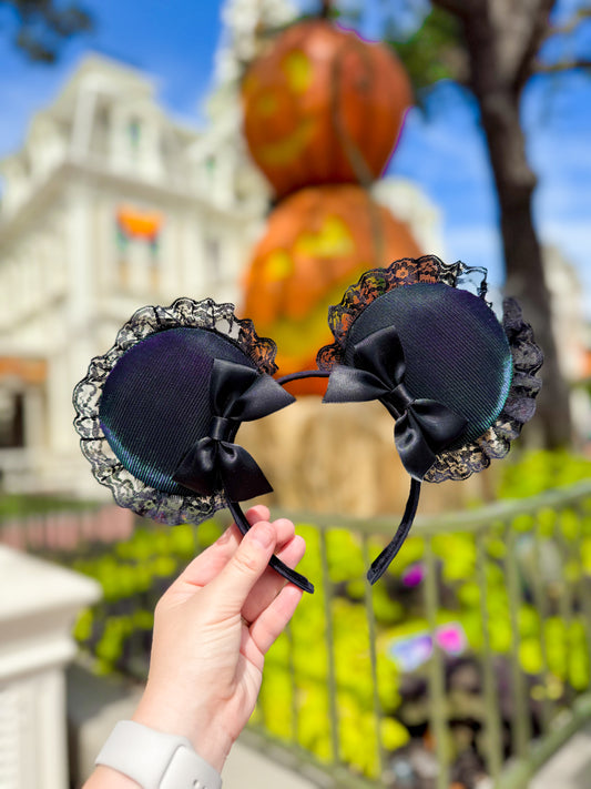 Black decorative mouse ears with bow and lace details held in front of a Halloween-themed background.