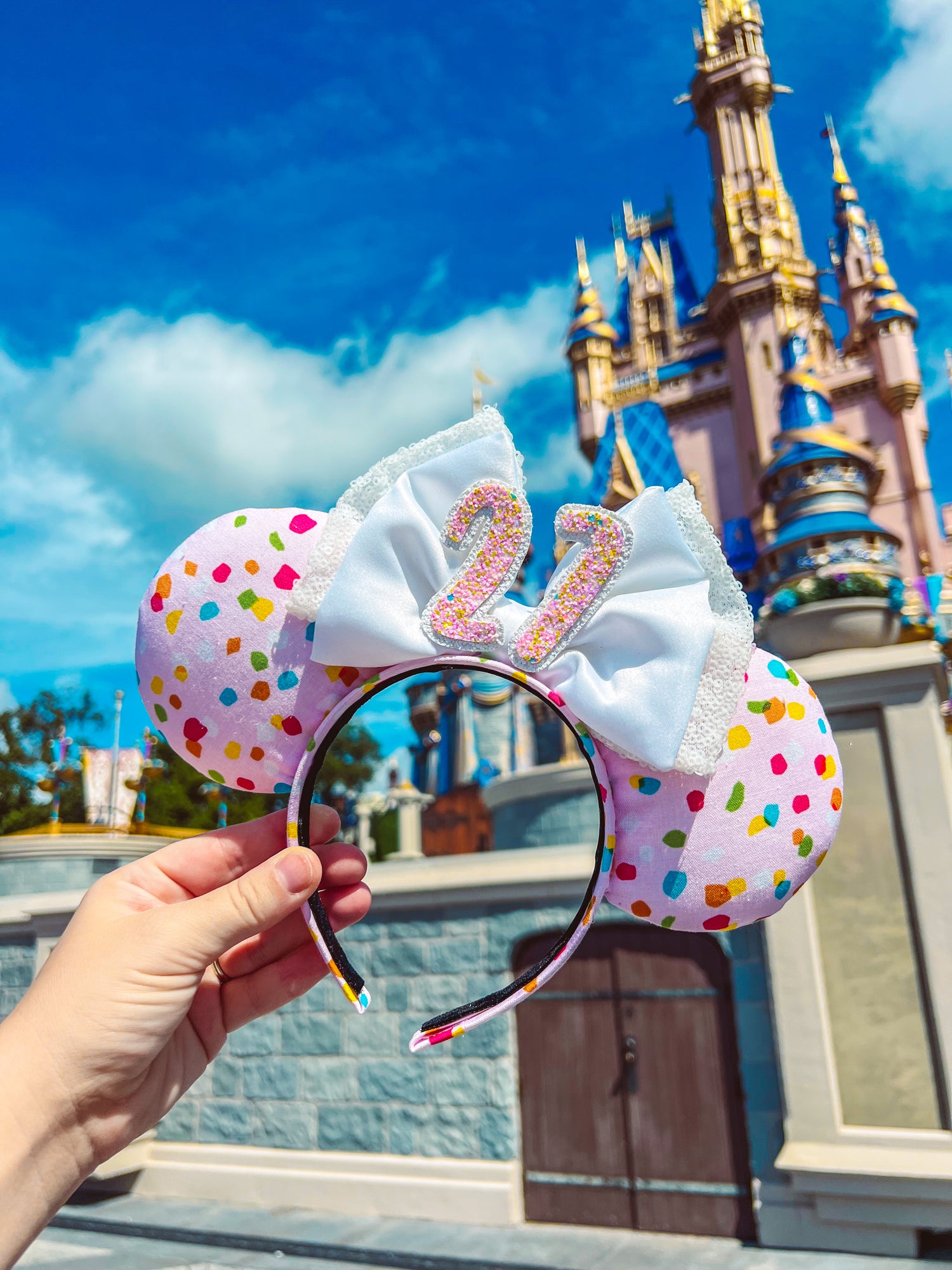 A hand holding a pair of birthday mouse ears with a multicolored polka dot pattern and a double white satin bow in front of a castle.