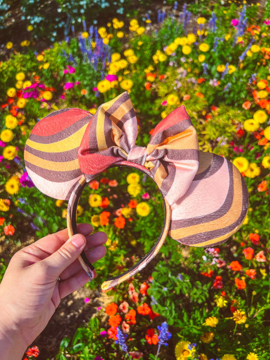 A hand holding a pair of groovy striped mouse ears in orange, yellow, and pink colors, with a background of a colorful floral garden.